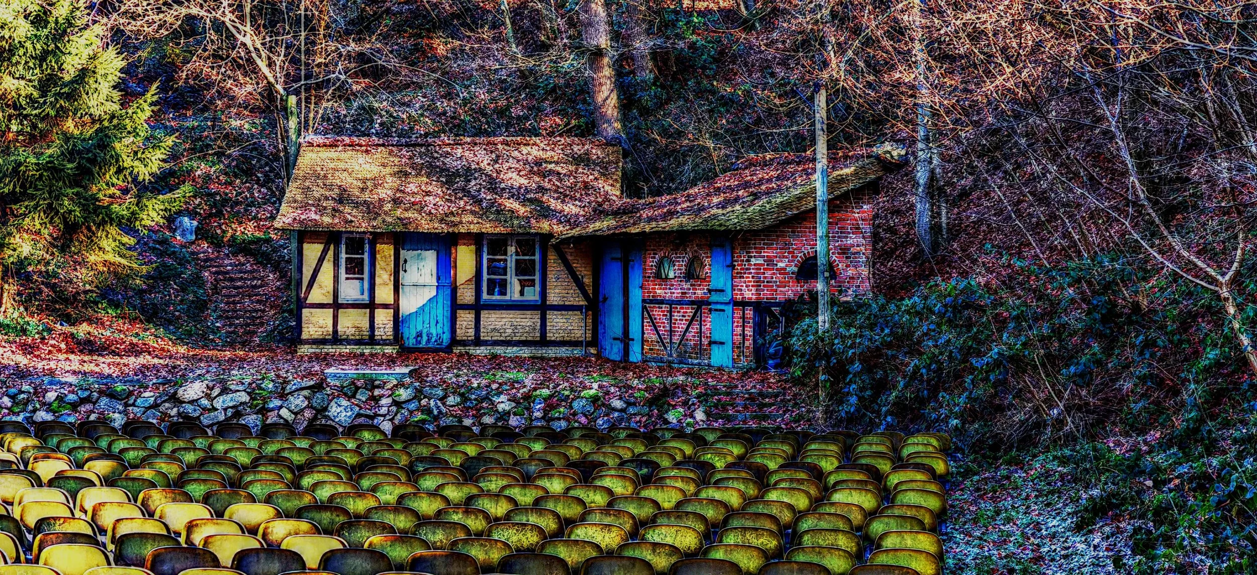 Ein verfallenes Haus im Wald, mit bunten Bäumen und herabfallenden Blättern, im Vordergrund sind leere, gelbe und schwarze Stühle aufgestellt.