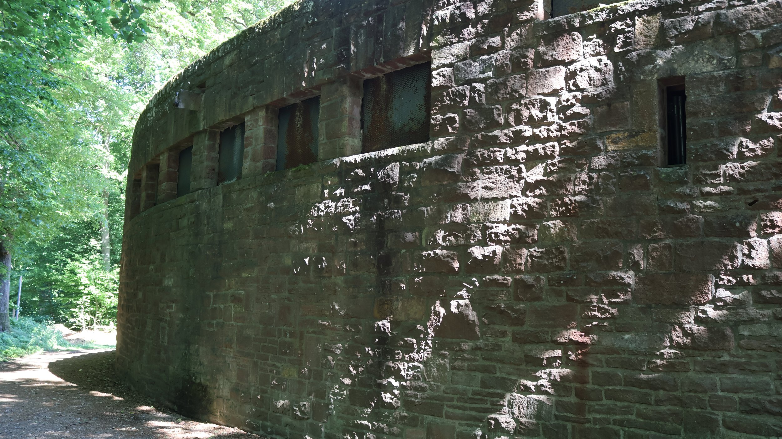 Ein Rundbau aus alten Steinen, umgeben von Bäumen und Schatten, mit kleinen rechteckigen Fenstern.
