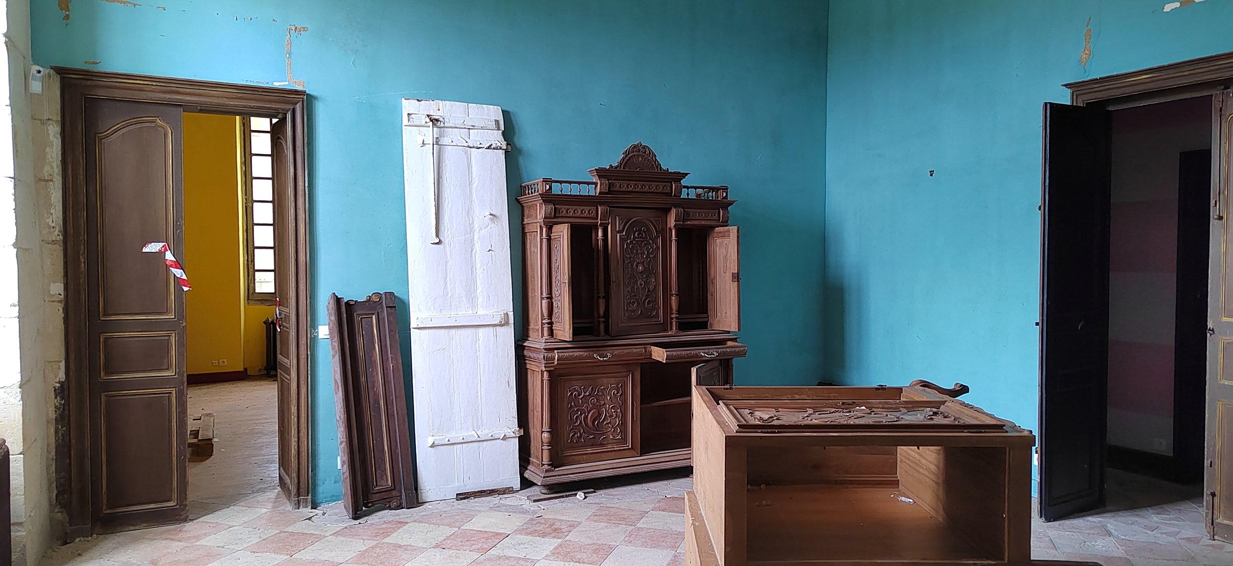Innenraum eines alten, unvollendeten Raumes mit blauer Wandfarbe, Holzmobiliar und offenen Türen. Es stehen verschiedene Holzmöbel, darunter ein großer Schrank mit kunstvollen Schnitzereien und eine niedrigere Kommode sowie ein unvollständiger Tisch 