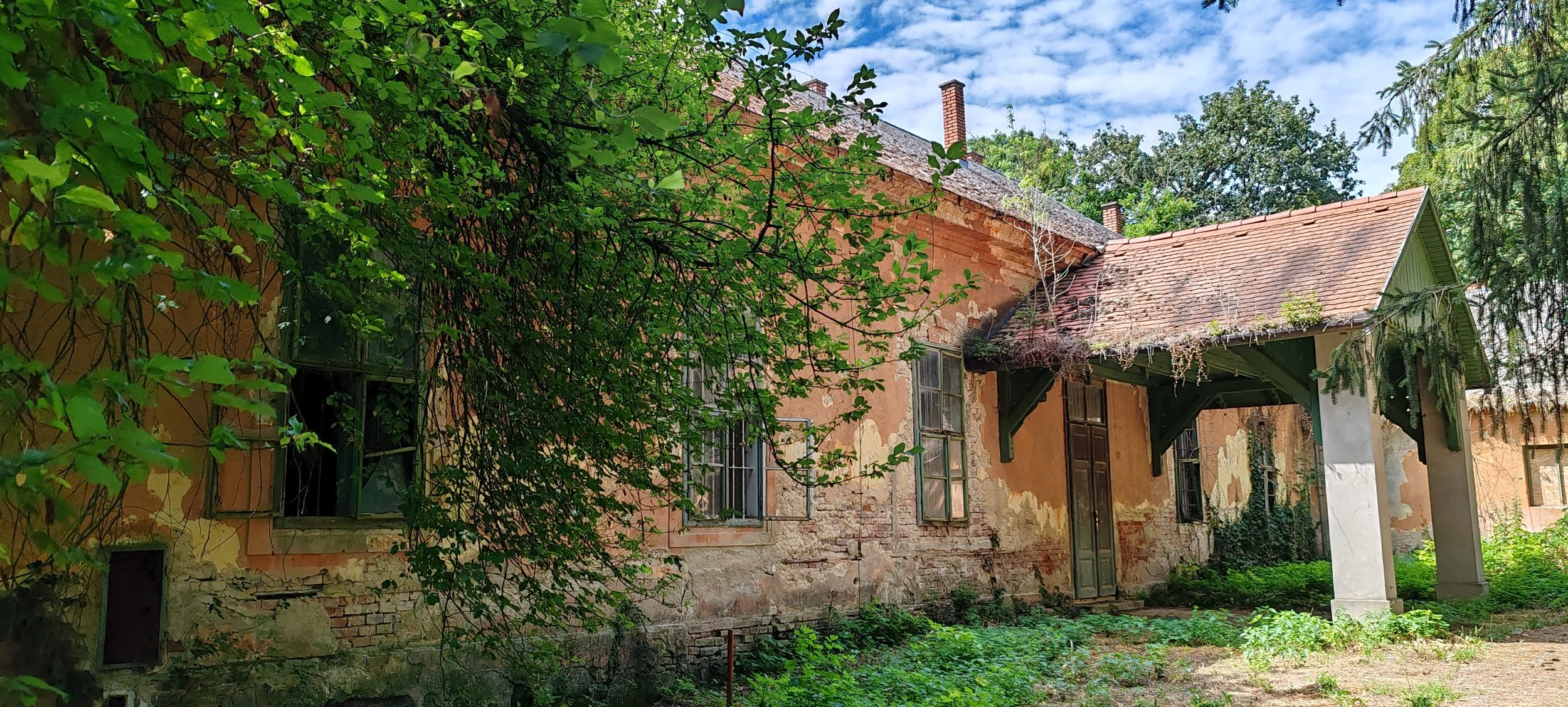 Verfallenes Haus mit bröckelnder Putzfassade, mehrere offene Fenster, und einem großen Vordach, umgeben von grünen Büschen und Bäumen unter einem blauen Himmel.