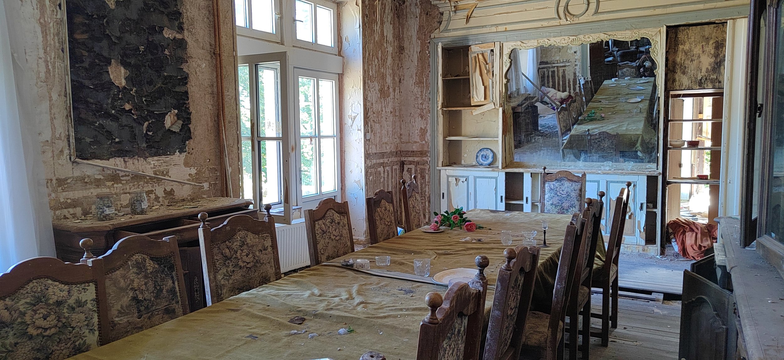 Ein alt aussehender Speisesaal mit einem langen Esstisch, umgeben von Stühlen, und einem großen Spiegel an der Wand, während die Wände abblättern und das Zimmer renovierungsbedürftig ist.