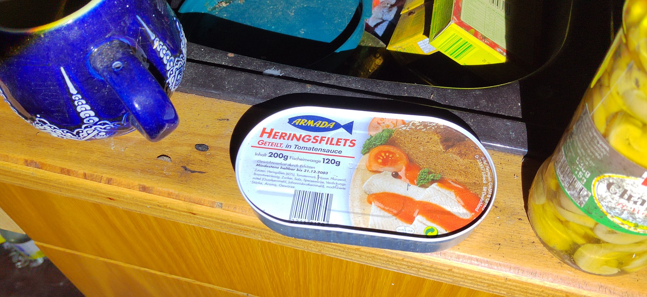 Dosen mit Heringsfilets in Tomatensoße, daneben eine blaue Kaffeekanne und ein Glas mit eingelegten Gewürzgurken