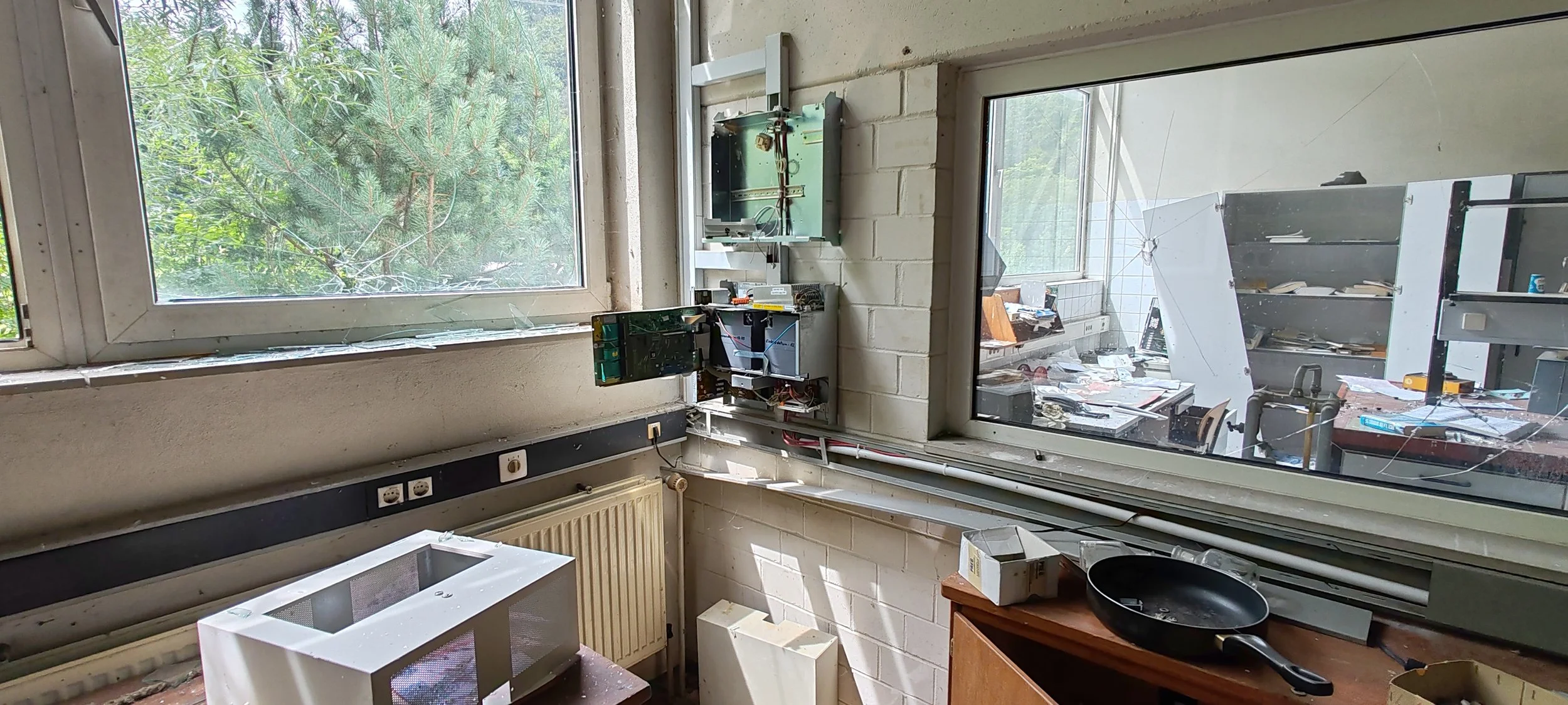 Innenraum eines zerstörten Raumes mit beschädigten Fenstern, verstreuten Papieren und zerbrochenen Gegenständen, Blick durch das Fenster auf den Außenbereich mit Bäumen.