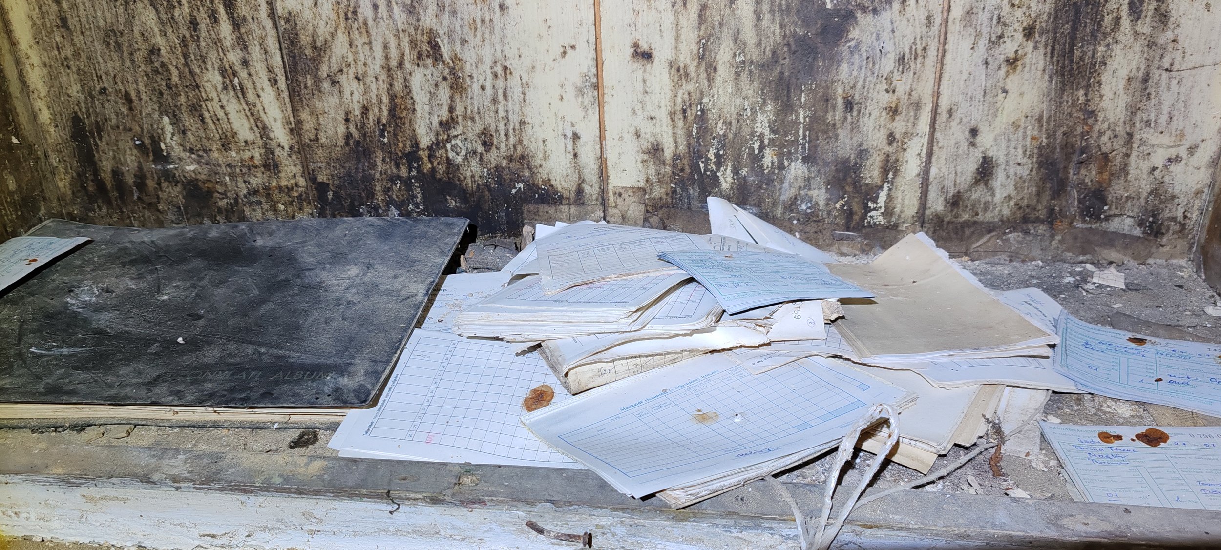 Datenblätter und Dokumente liegen ungeordnet auf einer verstaubten Fensterbank, dahinter ist eine schmutzige, rostige Wand.