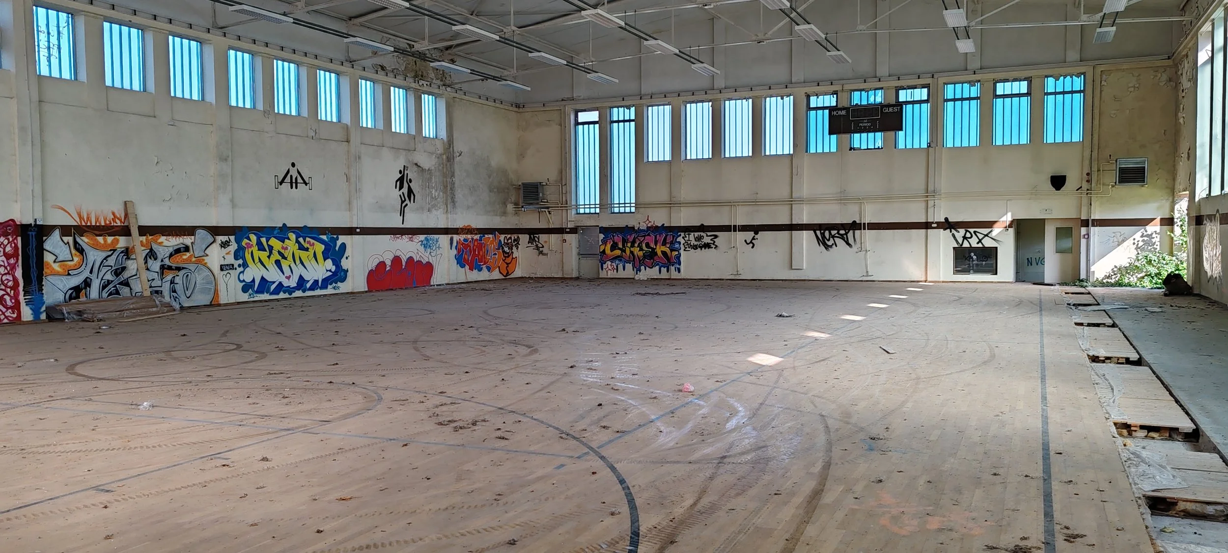 Leeres, verfallenes Hallenbad mit Graffiti an den Wänden, zerbrochener Bodenbelag auf der rechten Seite, große Fenster oben lassen natürliches Licht herein.