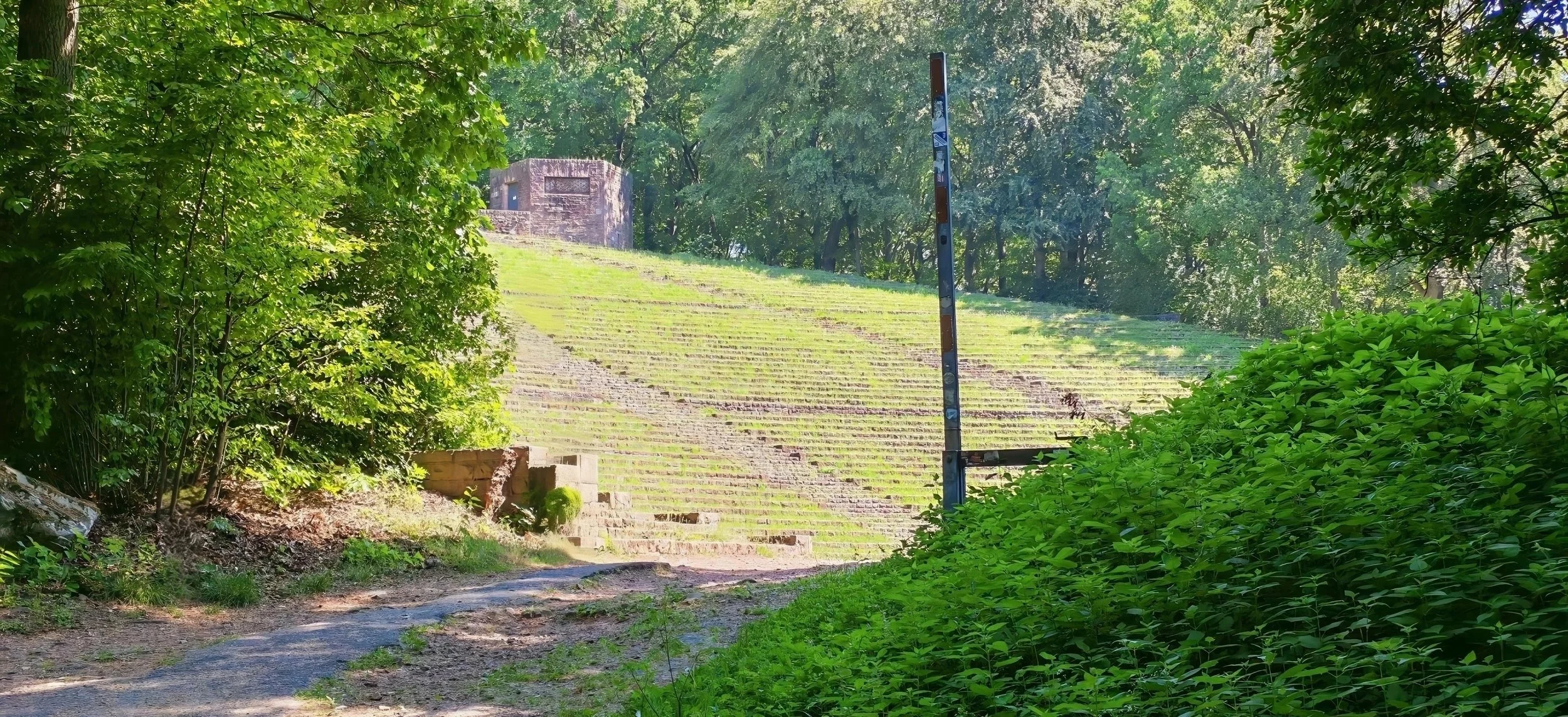 Ein grüner, bewaldeter Hang mit einem Weg im Vordergrund und einer kleinen Abrissstelle, im Hintergrund ein alter Bunker auf einer Wiese.