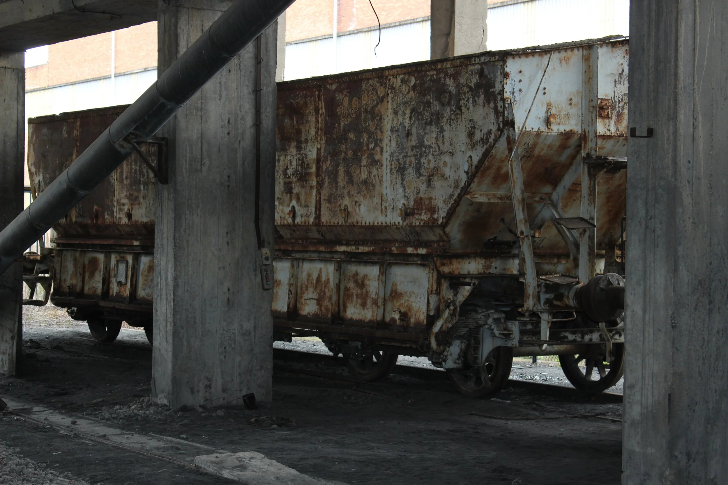 Verrosteter, alter Eisenbahnwagen unter einem Betonüberbau, mit großen schwarzen Rohrleitungen darüber.