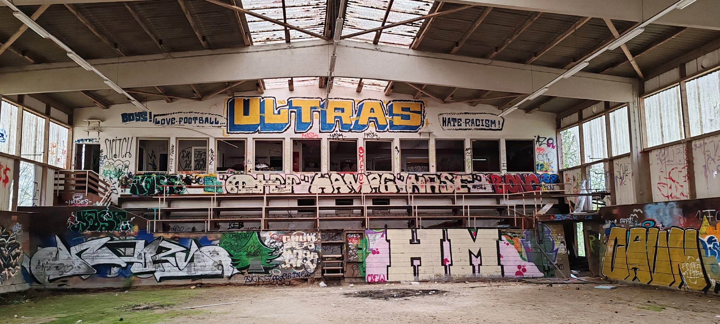 Innenansicht eines verfallenen Gebäudes mit Graffiti an den Wänden, im oberen Bereich steht 'ULTRAS' in großen blauen Buchstaben, umgeben von Worten wie 'Love Football!' und 'Hate Racism'.