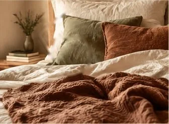 Unmade bed with cream and rust linens, a sage green pillow, and a small plant and books on the nightstand