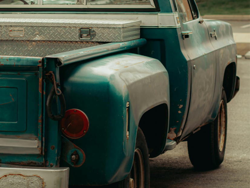 Close-up of a vintage blue pickup truck with rust, dents, and a diamond-plate tool box in the truck bed.