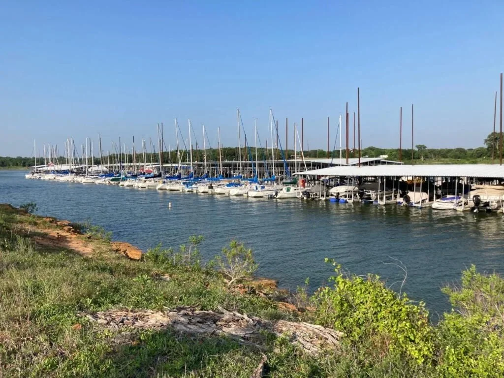 Marina with sailboats docked along a pier on a calm body of water under a clear blue sky, with greenery and rocks in the foreground.