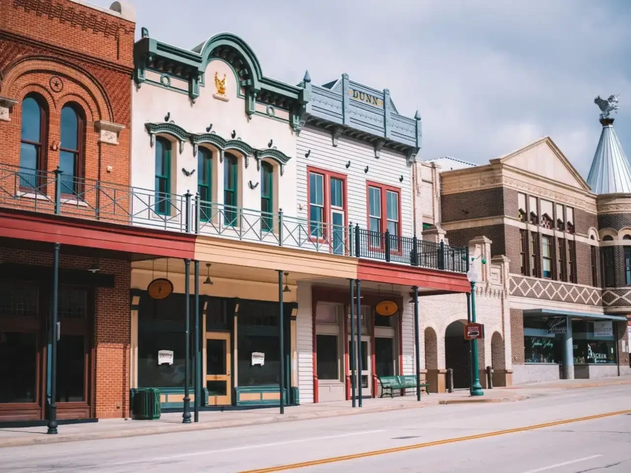 Empty street with historic-style buildings with colorful facades, storefronts, and empty sidewalk with benches and street lamps under cloudy sky.