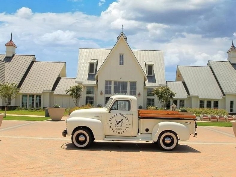 A vintage white pickup truck parked on a paved area in front of a large white church with a steep roof, surrounded by small trees and large planters, under a partly cloudy sky in Northlake, TX—a growing hub for dustless blasting.