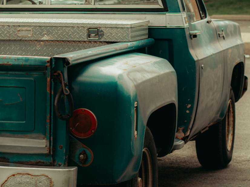 Close-up of an old, weathered teal pickup truck with rust spots, a metal toolbox in the truck bed, and some rust around the tail light.