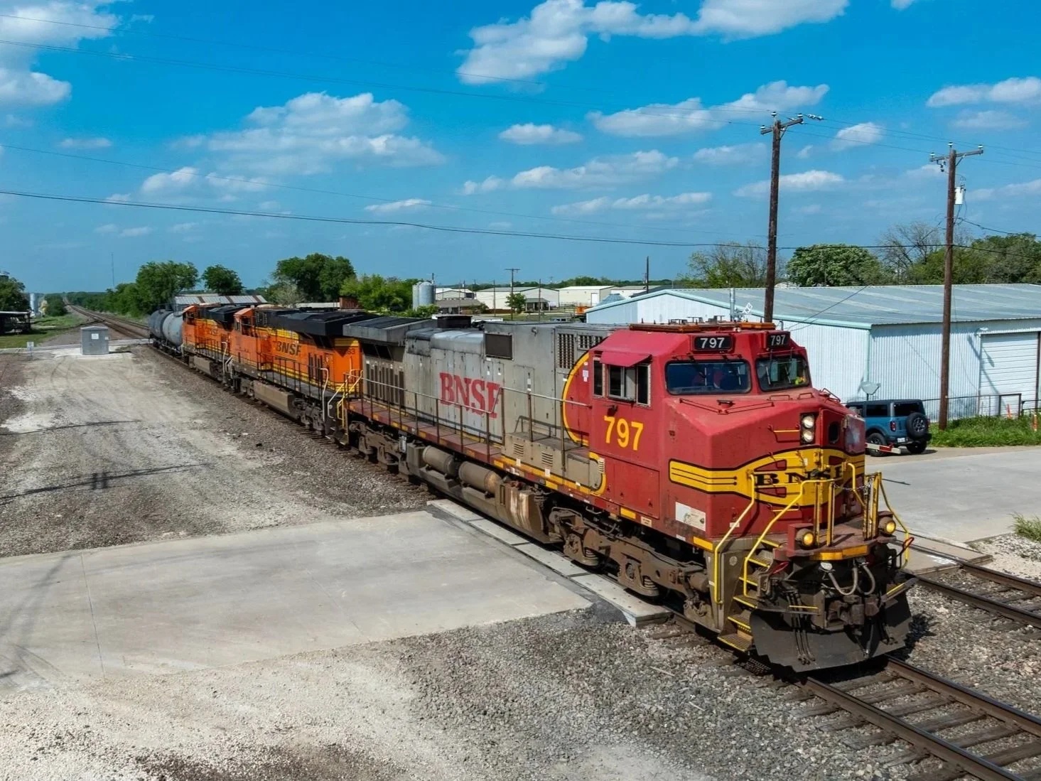 A BNSF freight train with orange and gray locomotives moving along the tracks near a small station with warehouses and electrical poles under a partly cloudy blue sky outside Justin, Texas.