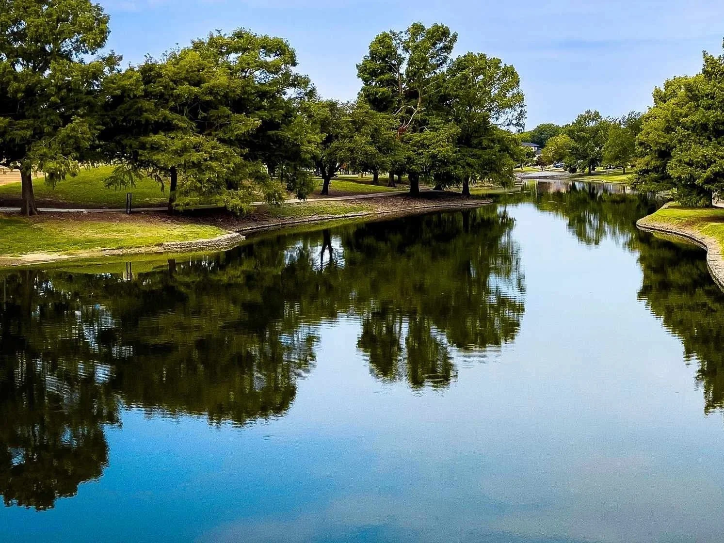 A peaceful park with a calm, reflective body of water lined with green trees and grass on a sunny day in Farmers Branch