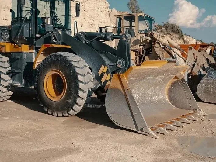 A large front loader construction vehicle parked on dirt ground with a hill and partly cloudy sky in the background.