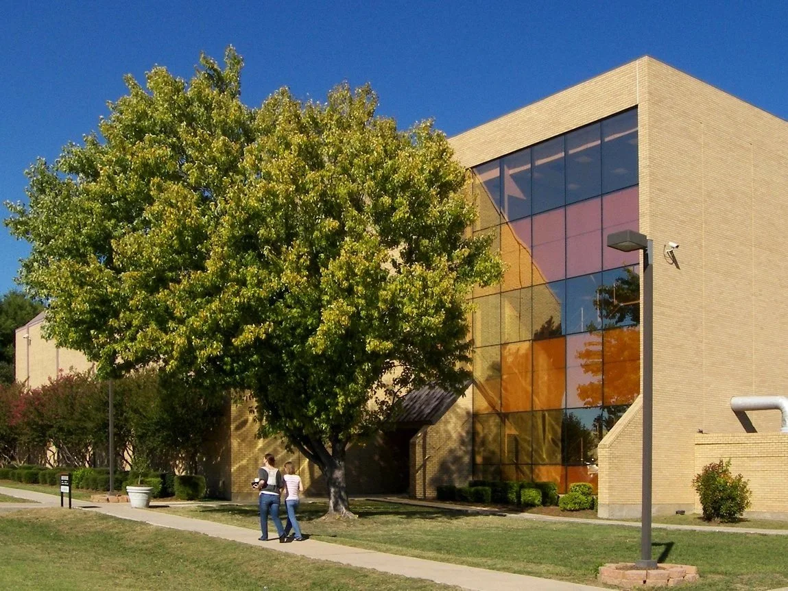 A modern brick building with a large glass windowed facade reflecting the trees and sky outside, situated on a well-maintained lawn with two people walking on a sidewalk nearby, during daytime with clear skies. Dallas Community college .