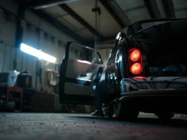 A dark vintage car with tail lights on inside a garage, with an open driver-side door and a person standing next to it.