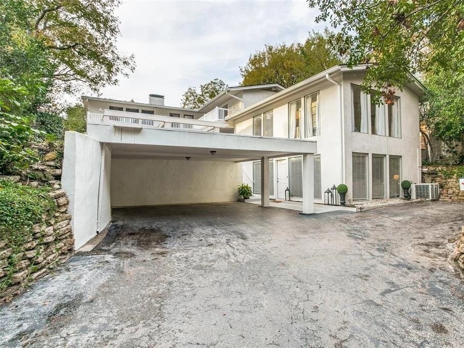 Modern white two-story house with a driveway and covered parking space beneath the upper level, surrounded by trees and greenery, with a stone wall on the left side.