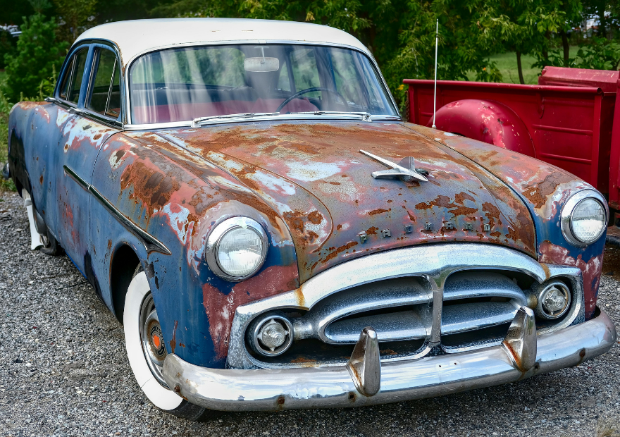 An old, rusted vintage car with a white roof and faded blue and rust-colored body, parked outdoors near a red trailer, with a background of green trees.