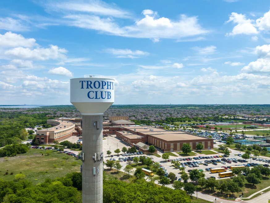 A tall water tower with a round top displaying 'TROPHY CLUB' in large blue letters, surrounded by a large school or community center with parking lots, trees, tennis courts, and a vast neighborhood under a partly cloudy sky.