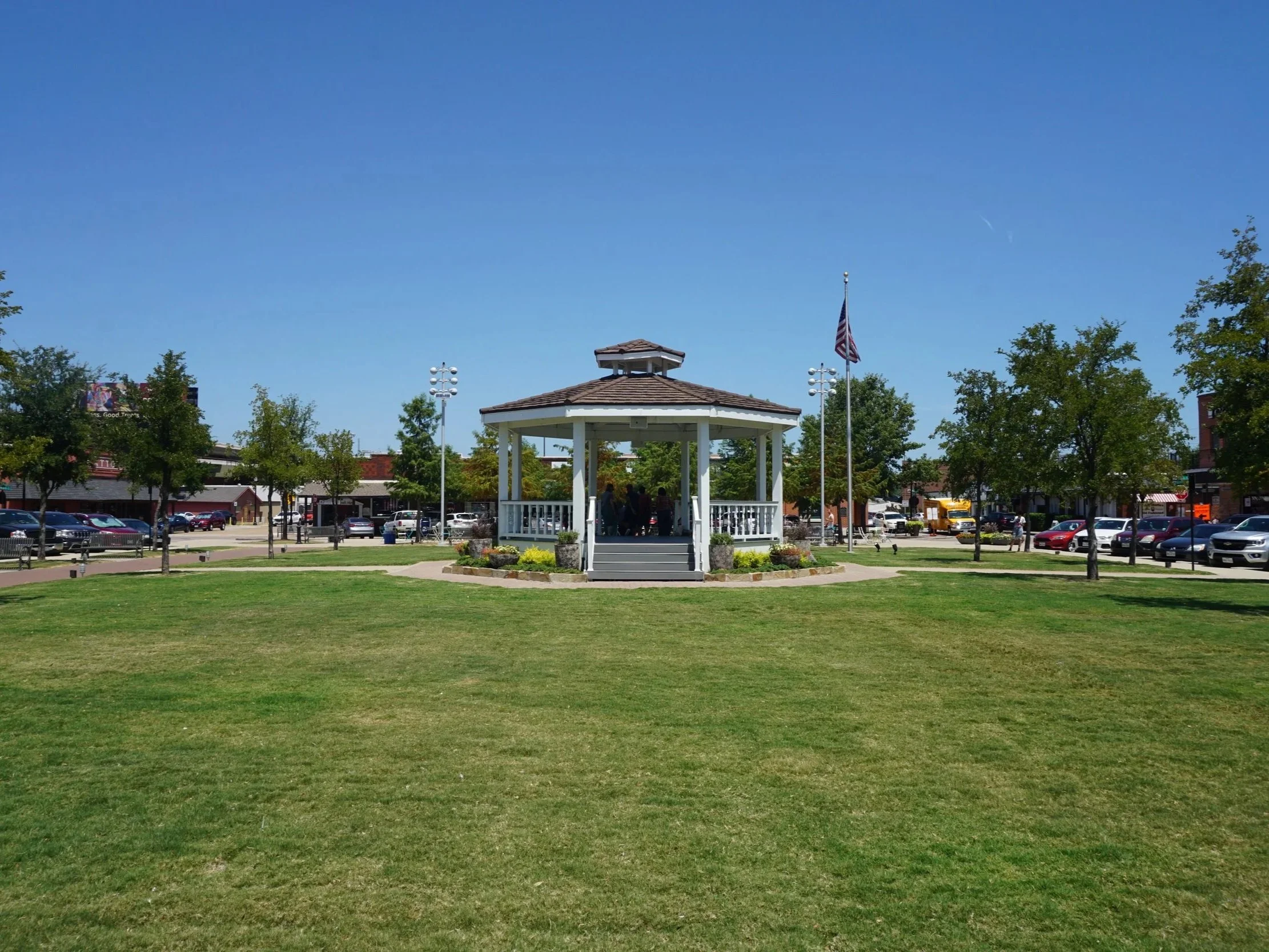 A park with a green lawn, trees, a white gazebo, and an American flag on a clear, sunny day.