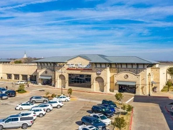 A large shopping center with a parking lot in front, filled with cars, under a blue sky with some clouds.
