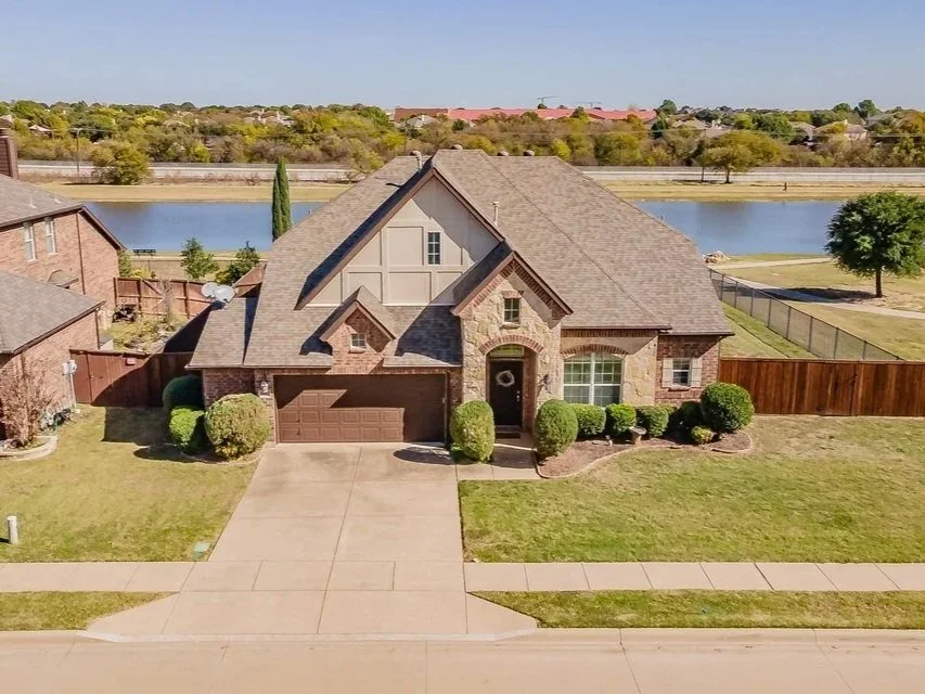 A suburban house with a brick and stone exterior, front yard with bushes, driveway, and a backyard lake view. The type of property that would benefit from a driveway cleaning before being listed for sale.