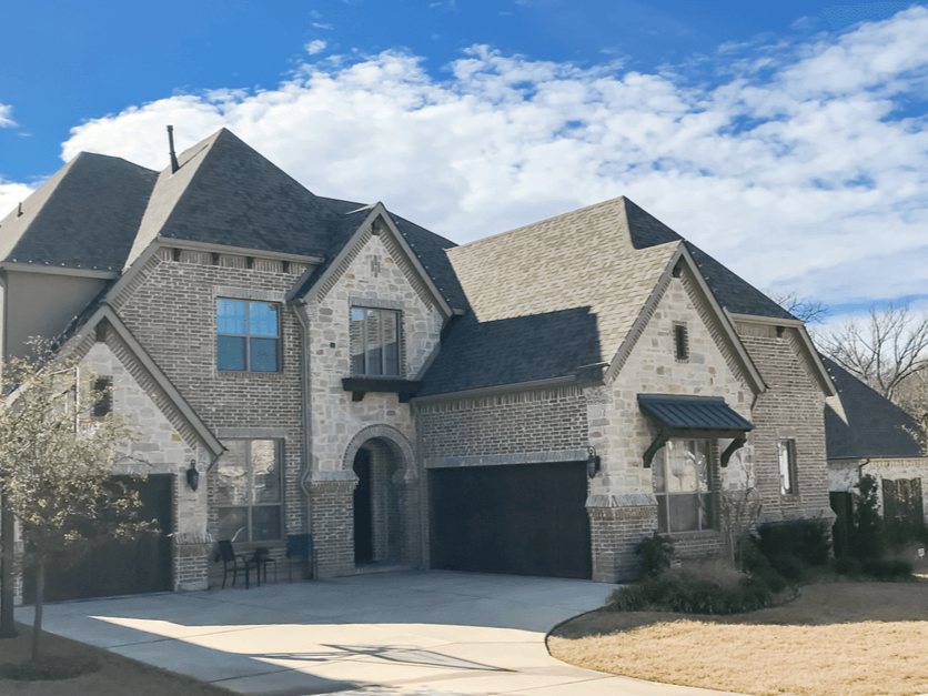Front view of a large two-story house with a mixed brick and stone facade, multiple gabled roofs, a double garage, and a small front porch under a blue sky with scattered clouds.