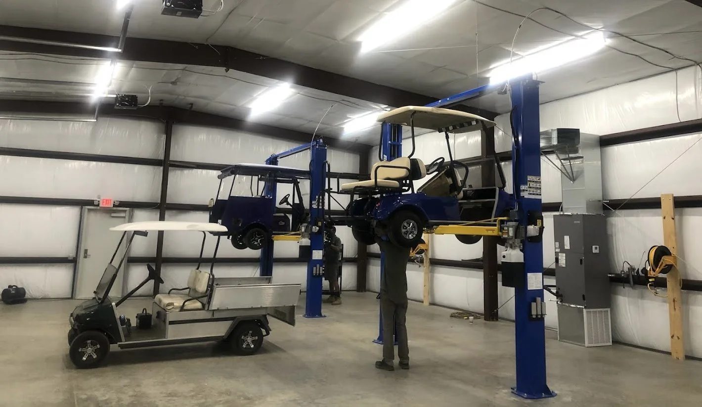Inside a large industrial garage with a hydraulic lift holding two golf carts, one of which has a cream-colored seat. A person is working underneath one of the golf carts, and a small electric golf cart is parked below the lift.