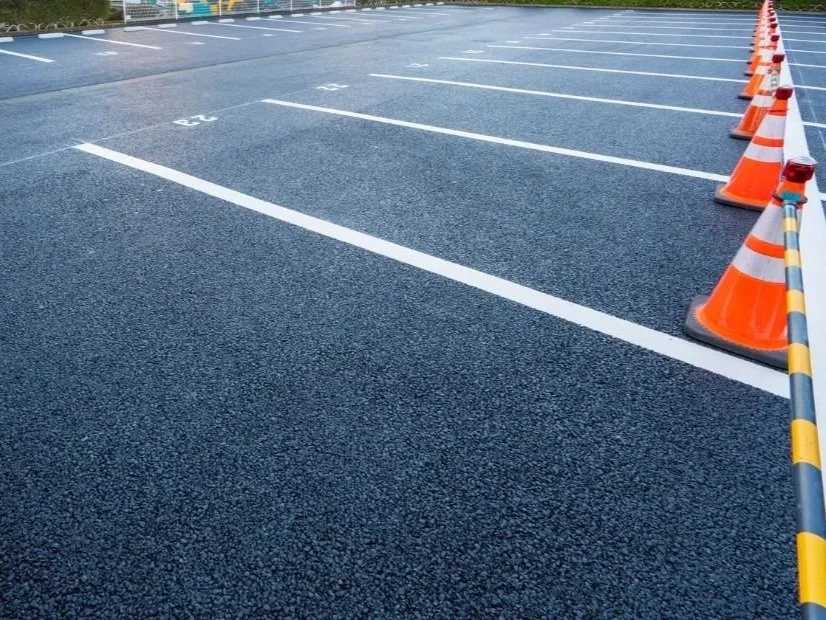 Empty parking lot with newly painted parking spaces, orange traffic cones along the side for construction or maintenance, and a striped barrier.