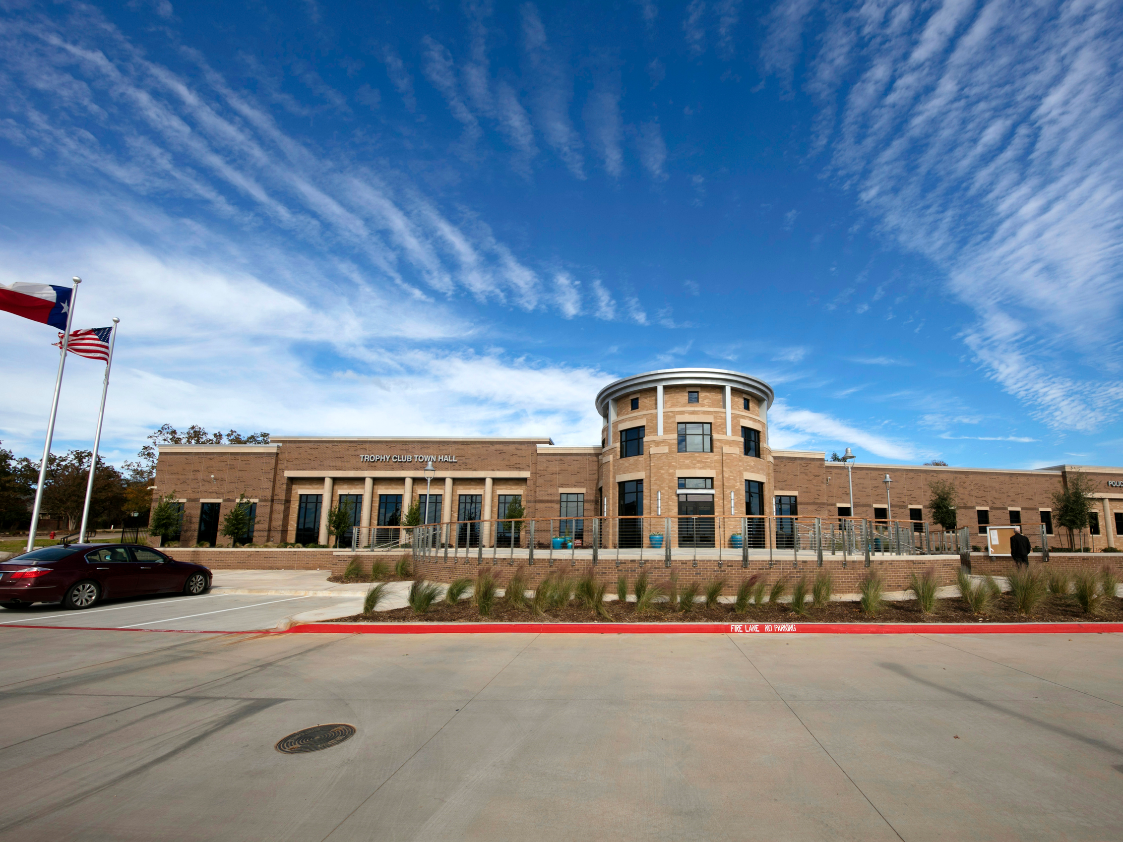 Front view of Trophy Club Town Hall building under blue sky with scattered clouds, flags flying on flagpoles, and a parking lot in the foreground.
