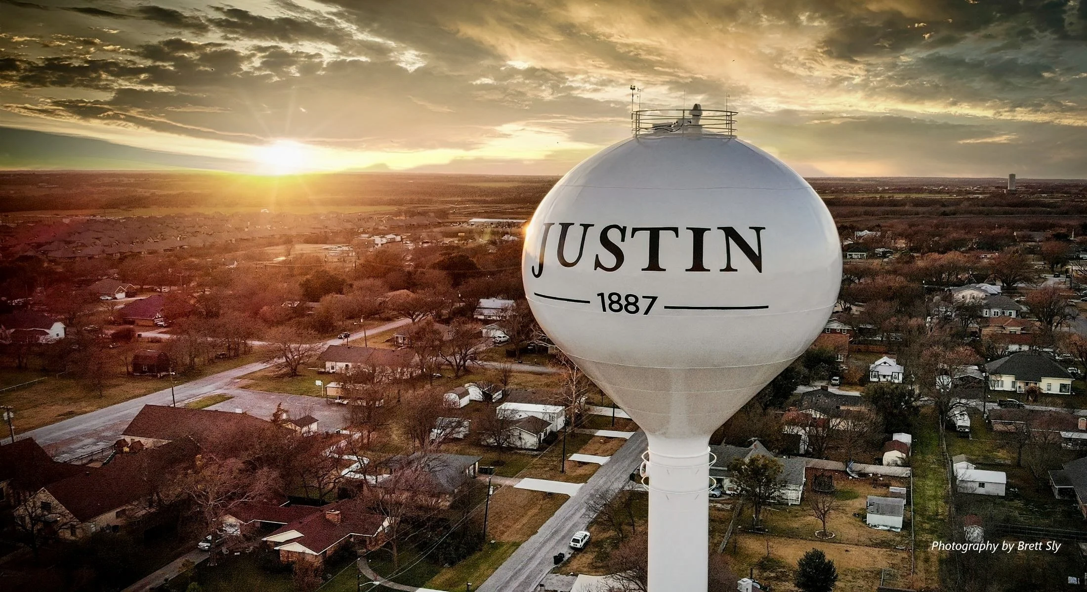 A water tower with the label 'Justin 1887' stands above a residential neighborhood during sunset with an orange and yellow sky, trees, streets, and houses below.