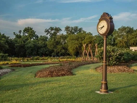 A landscaped park with a vintage-style clock on a tall post, surrounded by green grass, trees, and garden beds under a partly cloudy sky in Famers Branch,