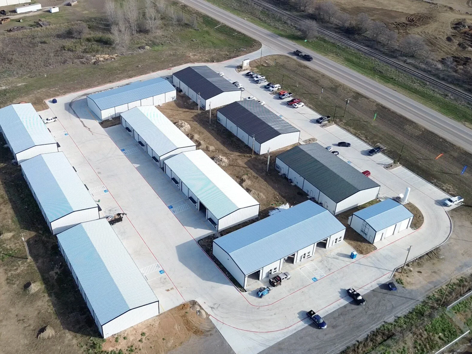 Aerial view of a new commercial or industrial complex with multiple white metal buildings, parking lots, and cars, situated near a highway.