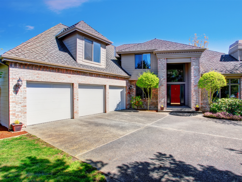 A modern two-story house with a brick and siding exterior, three-car garage doors, and a paved driveway. Front yard with potted plants, small trees, and shrubs under a clear blue sky.