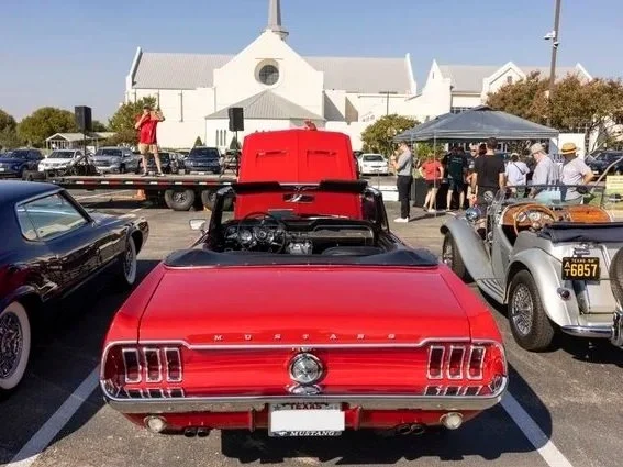 Rear view of a red vintage Ford Mustang convertible at a car show, with other classic cars and people in the background near a white church building.