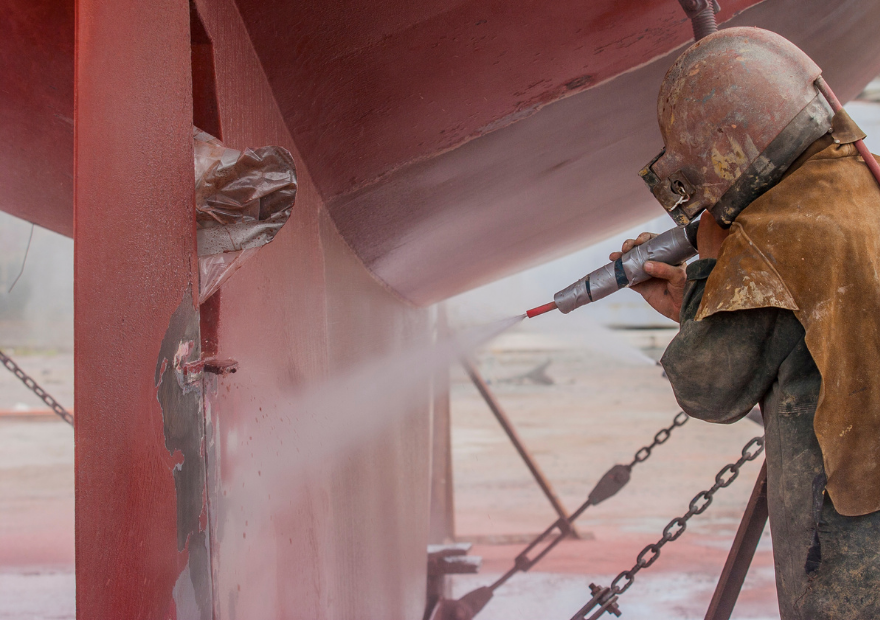 A worker in protective gear is wet blastinga large red ship's hull at a shipyard.