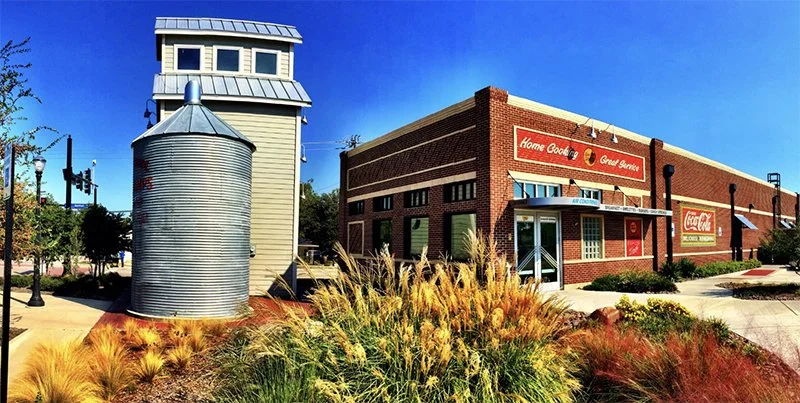 A vintage-style brick building with a sign indicating a home goods and general service store, with Coca-Cola signage. In front, there is a grassy area with ornamental weeds or grasses, a metal grain silo-like structure, and sidewalk lighting. The sky is clear and blue.