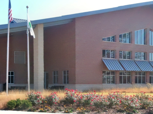 A large modern brick building with a sloped metal roof over the windows, a flagpole with flags, and landscaped flower beds in front.