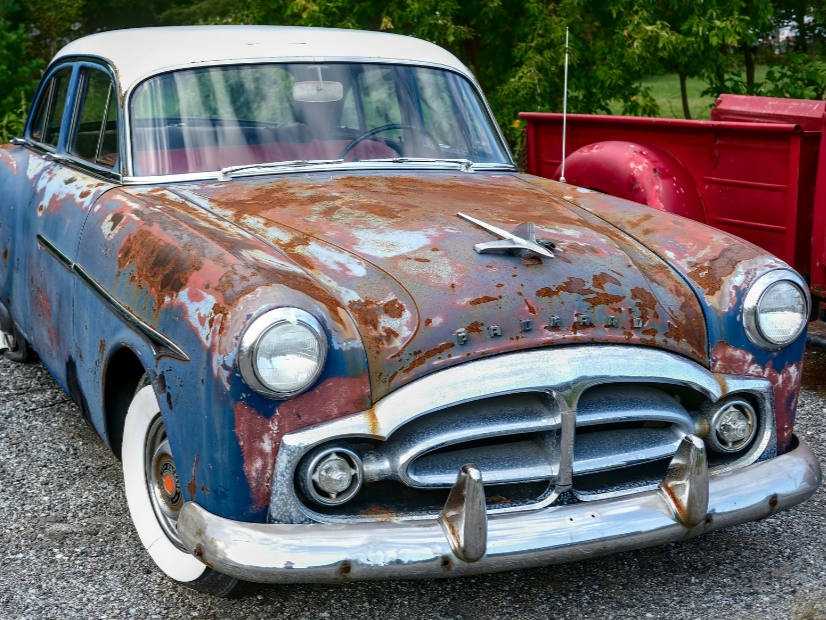 An old, rusty vintage car with a white roof and front grille, parked outdoors on gravel with green trees in the background.