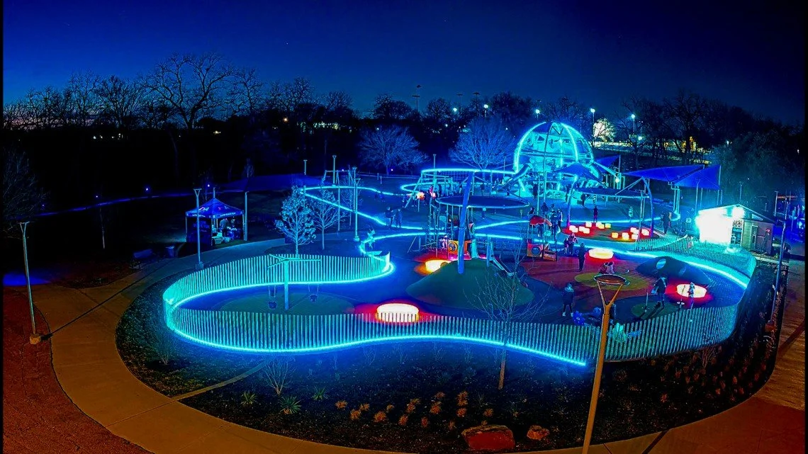 Nighttime view of illuminated outdoor playground with neon blue lights, climbing structures, slides, and children playing in Farmers Branch Texas