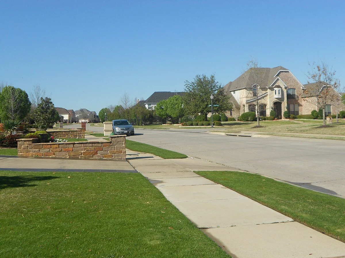Residential neighborhood street with sidewalks, cars parked, well-maintained lawns, and large houses under a clear blue sky.
