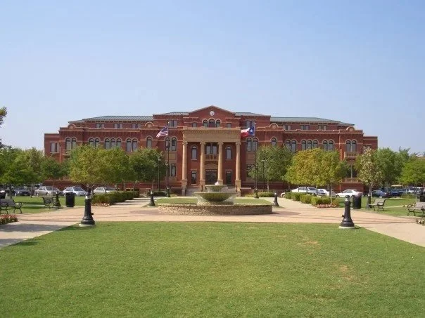 Large brick building with flags, surrounded by trees, with a fountain and lawn in front.