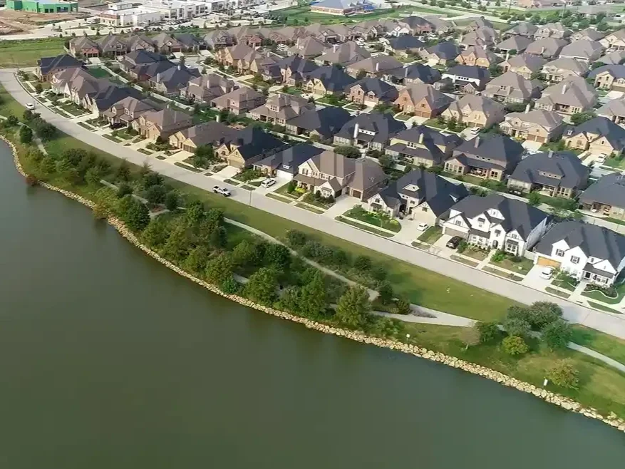 Aerial view of suburban houses along a waterfront with a walking path and greenery.