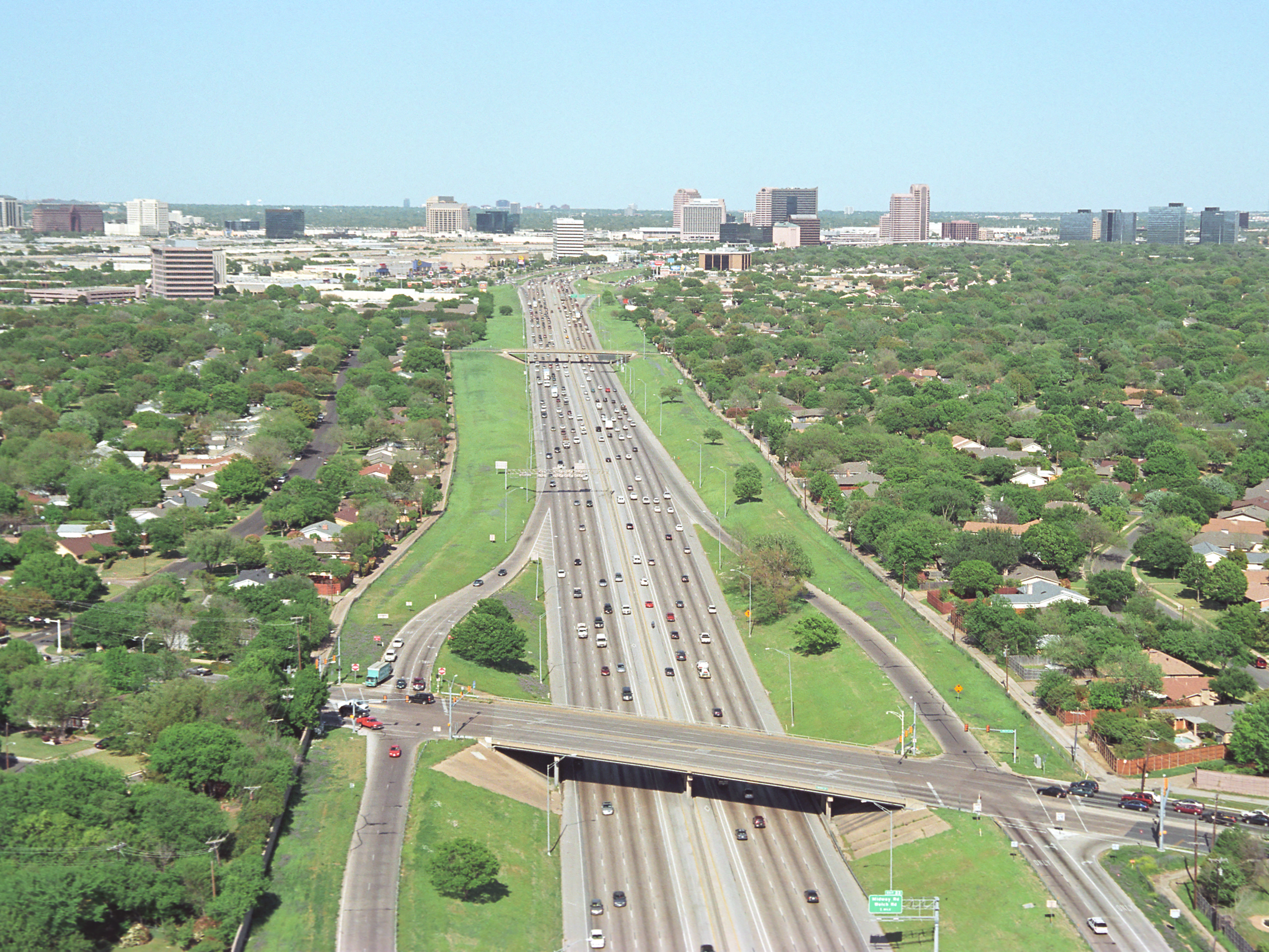 An aerial view of a busy highway passing through a green city area with residential neighborhoods, leading to a downtown skyline with tall buildings in the background. IH-635 before the expansion.