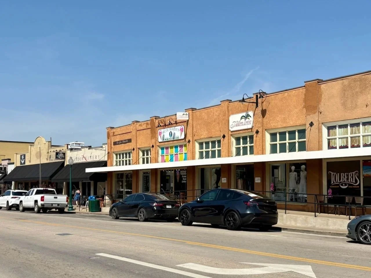 Street view of storefronts with parked cars in front, including businesses like Betti Maries, Palm Tree, and Tolbert's, under a clear blue sky.