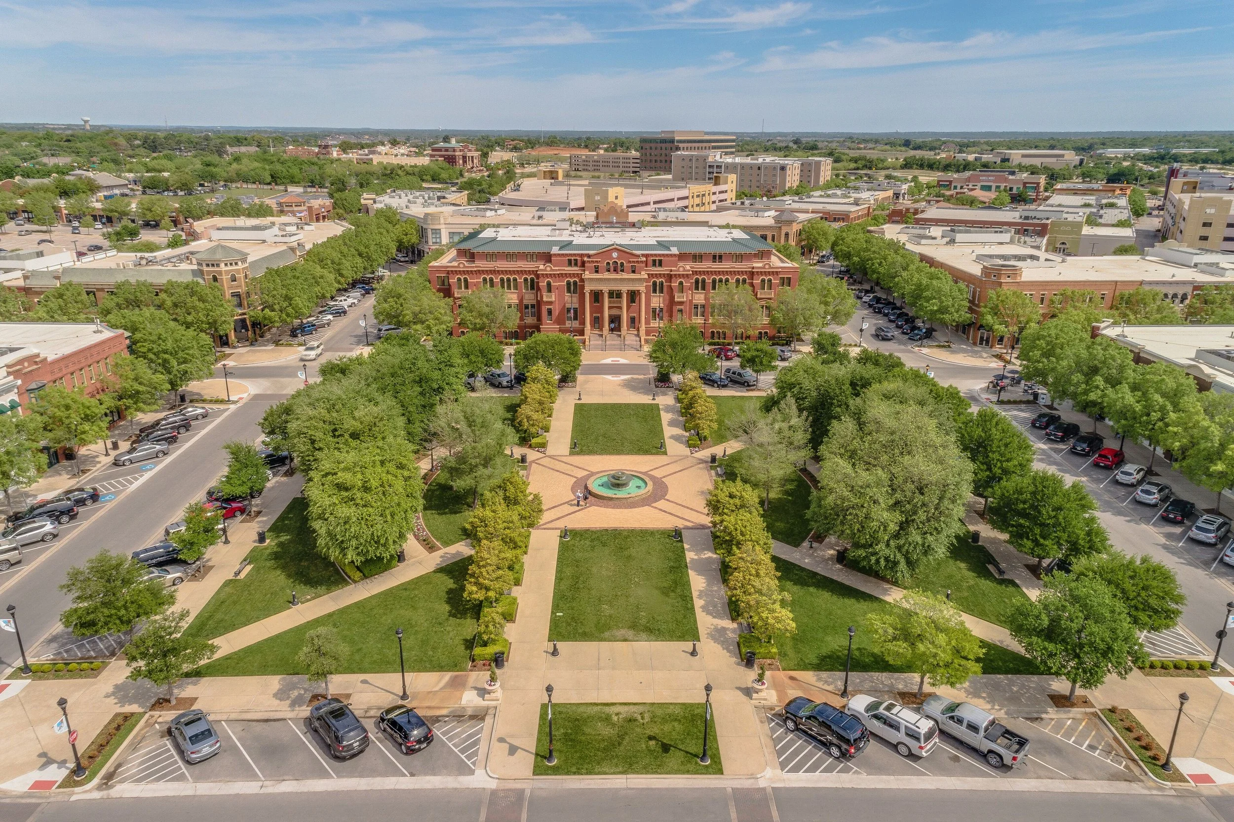Aerial view of a city square with a large red brick building, green trees, a fountain, and parking lots.