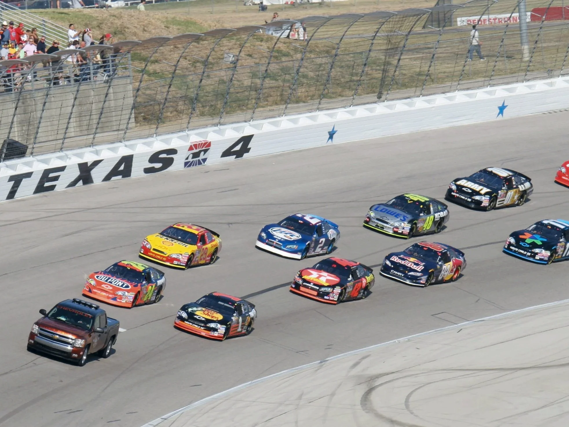Multiple stock cars racing on a track at Texas Motor Speedway, with fans in the stands and a sign that says 'TEXAS 4' on the wall.